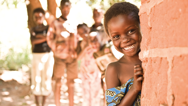 Mädchen in Uganda, schaut glücklich hinter einer Hausecke hervor. Bild: Oliver Rust