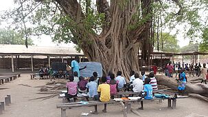 Eine kleine Schulklasse sitzt vor einem Baum im Freien und lässt sich von einem Lehrer an einer Schiefertafel etwas erklären. Bildung ist ein Schlüsselfaktor für eine friedliche Zukunft der südsudanesischen Bevölkerung. (Bild: Georg Rettenbacher, TF)