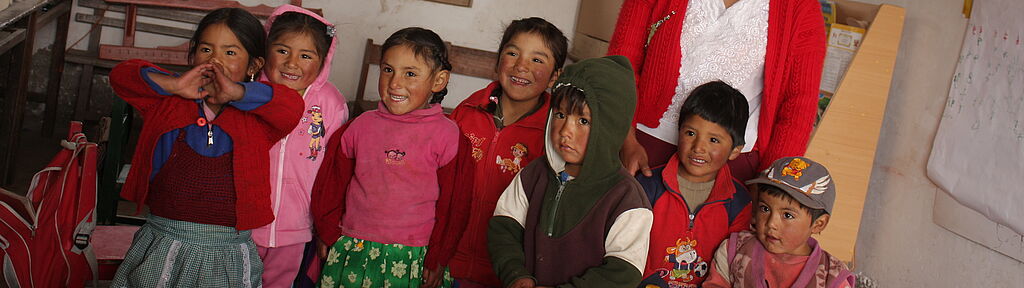 Peru: Eine junge Lehrerin steht mit einer farbig gekleideten und lachenden Kinderschar in einem Schulzimmer.