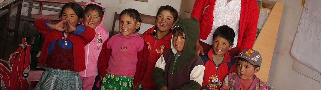 Sie befinden sich im Bereich Länder und Projekte: Peru Peru: Eine junge Lehrerin steht mit einer farbig gekleideten und lachenden Kinderschar in einem Schulzimmer.