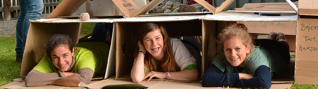 Veranstaltungskalender: Drei Teilnehmerinnen einer Nacht ohne Dach liegen in selbstgebauten Kartonhütten und lachen in die Kamera.
